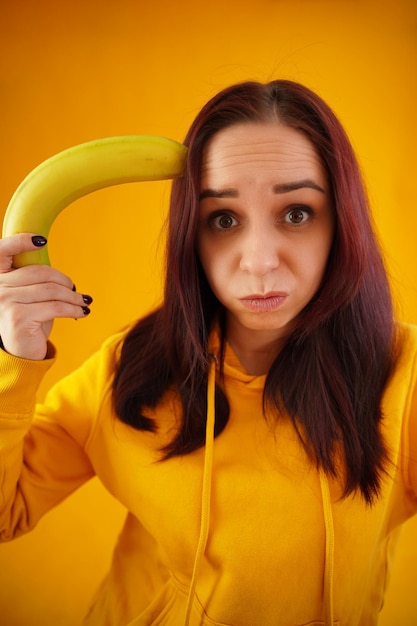 Retrato de jovem com banana em fundo amarelo Close-up de fêmea com capuz amarelo brinca com frutas imaginando-o como arma