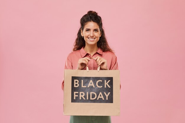 Retrato de jovem com cabelo encaracolado sorrindo enquanto segura uma compra da black friday contra o fundo rosa