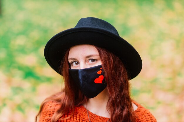 Retrato de jovem com camisola de malha vermelha, chapéu com uma máscara médica elegante preta no parque outono amarelo. moda, estilo de vida, quarentena, coronavírus.