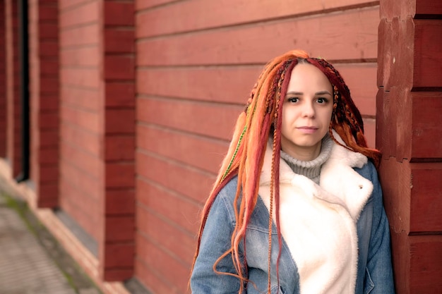 Retrato de jovem com dreadlocks de gengibre em pé encostado na parede na rua da cidade Mulher positiva olhando para a câmera sorrindo