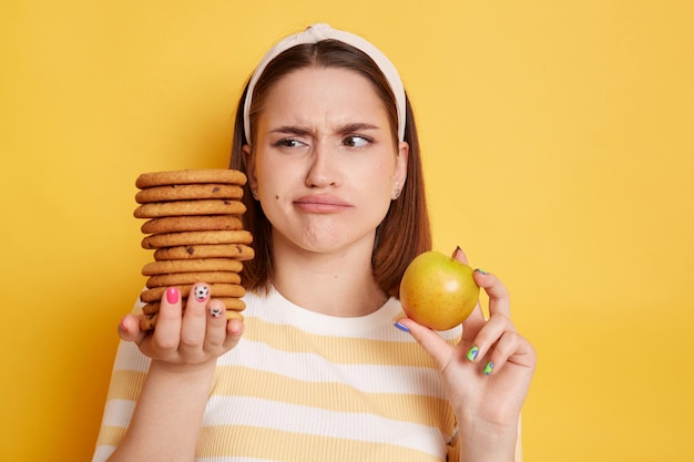 Retrato de jovem com lábios de beicinho vestindo camisa listrada e faixa de cabelo segurando biscoitos e maçã olhando carb com expressão confusa posando isolado sobre fundo amarelo