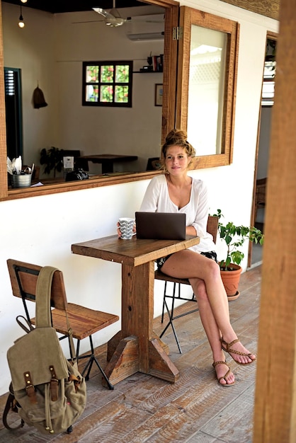 Retrato de jovem com laptop e caneca de café sentado na varanda de uma cafeteria