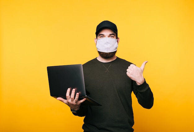 Retrato de jovem com máscara facial usando laptop e mostrando os polegares para longe