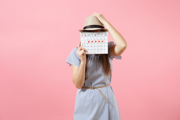 Retrato de jovem com vestido azul, chapéu segurando o calendário de períodos para verificar os dias de menstruação, isolados em um fundo rosa tendência brilhante. conceito médico, de saúde e ginecológico. copie o espaço.