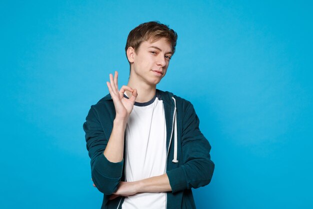 Retrato de jovem confiante em roupas casuais, em pé, mostrando o gesto de ok isolado em azul. Emoções sinceras de pessoas, conceito de estilo de vida.