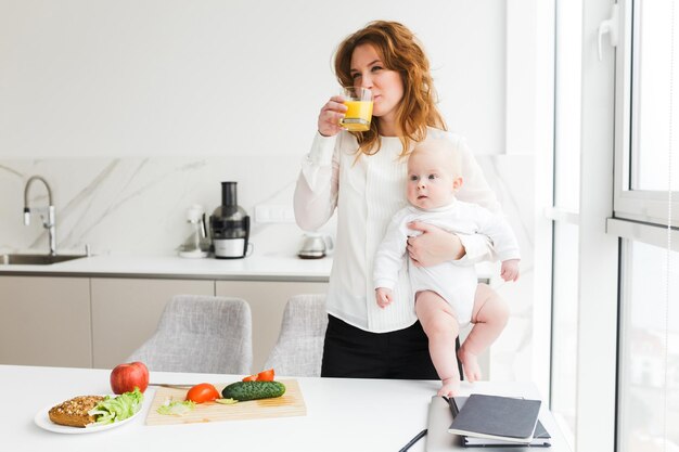 Retrato de jovem de pé e segurando seu bebê enquanto bebe suco de laranja e cozinha na cozinha