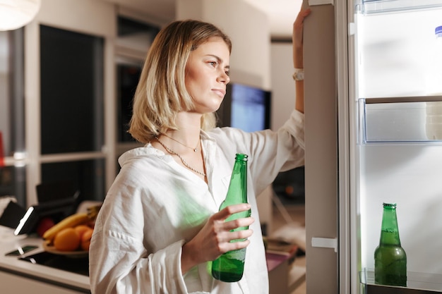 Retrato de jovem de pé na cozinha à noite e olhando pensativamente na geladeira aberta enquanto segura cerveja na mão em casa