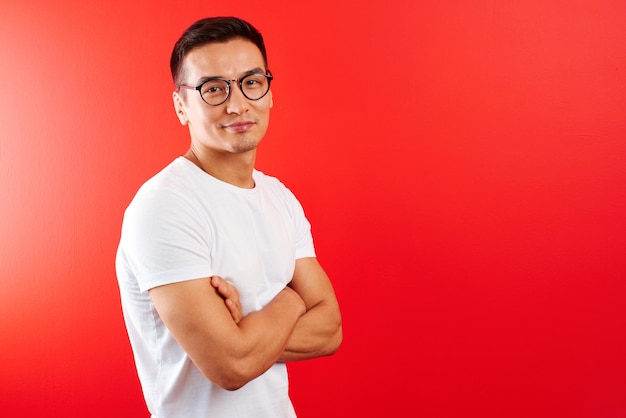 Retrato de jovem designer asiático vestido de casual e óculos contra a parede vermelha. Bonito homem cazaque sorri alegremente com as mãos cruzadas no estúdio