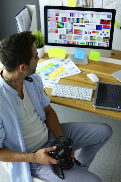 Retrato de jovem designer sentado no estúdio gráfico em frente ao laptop e computador enquanto trabalhava online