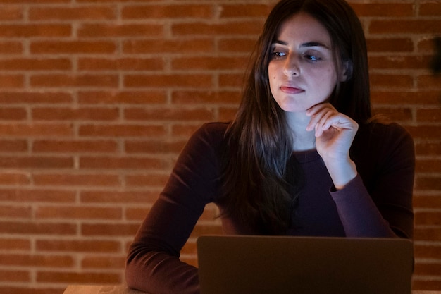 Retrato de jovem dúvidas mulher usando laptop à noite usando tecnologia moderna havinmg diversão com tela brilhante no escritório escuro ou em casa startup reunião de negócios on-line com mulher de negócios