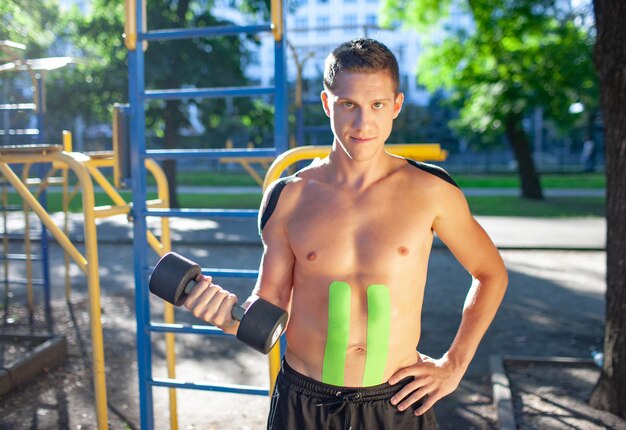 Retrato de jovem e bonito fisiculturista profissional caucasiano com fitas elásticas pretas e verdes no corpo carregando halteres no campo de esportes