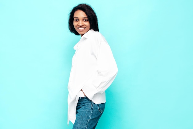 Retrato de jovem e linda mulher negra Modelo sorridente vestido com roupas de jeans de verão Mulher despreocupada sexy posando perto da parede azul no estúdio Bronzeado e alegre