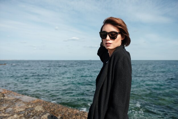 Retrato de jovem elegante com óculos escuros e casaco preto, caminhando perto do mar