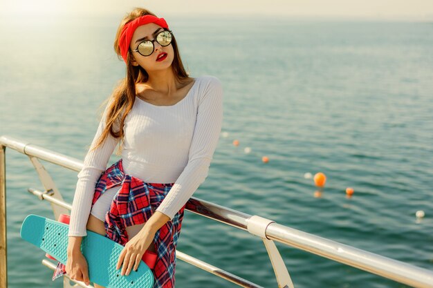 Retrato de jovem elegante em uma roupa da moda com o skate nas mãos, na praia contra o fundo do mar