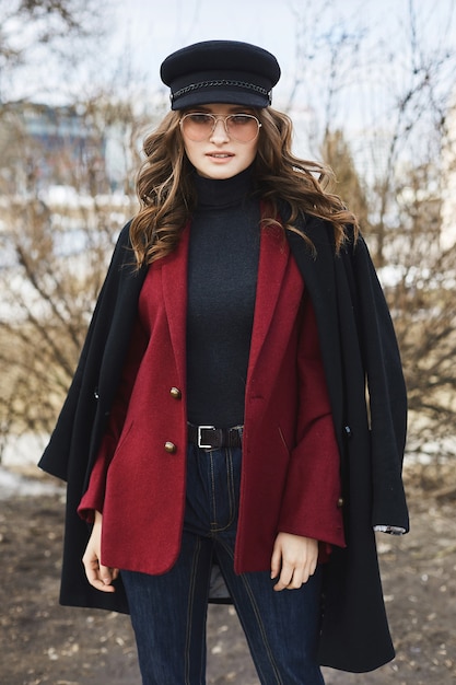 Retrato de jovem elegante usando óculos de sol da moda, casaco vermelho e chapéu posando ao ar livre.