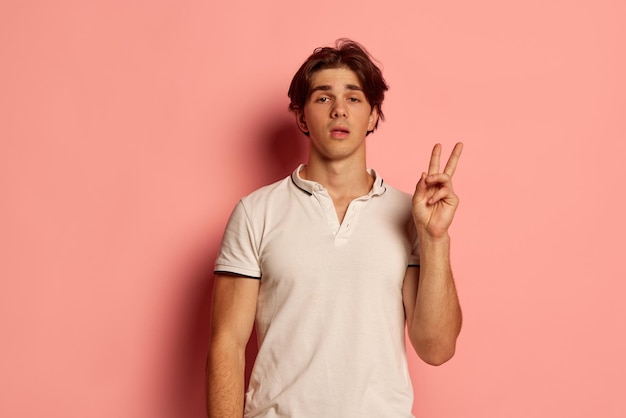 Retrato de jovem em camiseta branca casual posando com gesto de mão de paz sobre fundo rosa