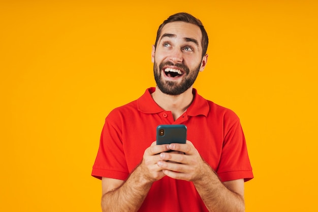 Retrato de jovem em camiseta vermelha sorrindo e segurando o smartphone isolado sobre o amarelo