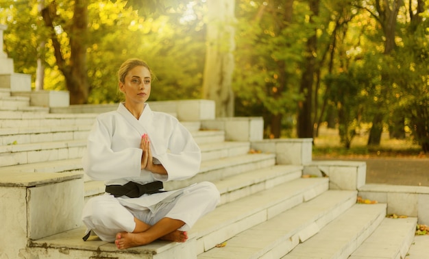 Retrato de jovem em quimono branco com faixa preta. mulher de esporte sentada na escada e meditando ao ar livre. artes marciais