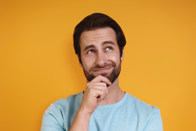 Retrato de jovem em roupas casuais, segurando a mão no queixo e desviando o olhar enquanto está de pé.