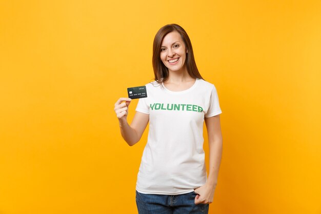 Retrato de jovem em t-shirt branca com voluntário de título verde de inscrição escrita segurar cartão de crédito isolado em fundo amarelo. Ajuda de assistência gratuita voluntária, conceito de trabalho de graça de caridade.