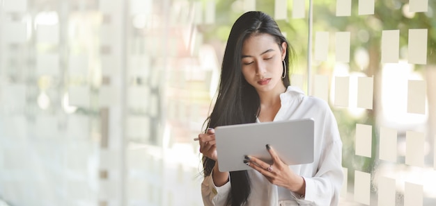 Retrato de jovem empresária asiática apaixonado usando tablet digital em pé no escritório moderno