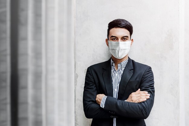 Retrato de jovem empresário asiático usando máscara cirúrgica e em pé na parede