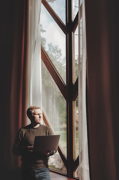 Retrato de jovem empresário com laptop na janela da sala de estar em casa de campo. Homem workaholic em roupas casuais em casa, trabalhando nas férias. Inspiração criativa e start-up de negócios. Copie o espaço