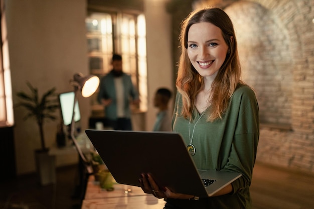 Retrato de jovem empresário sorridente segurando laptop e olhando para a câmera enquanto trabalhava à noite no escritório