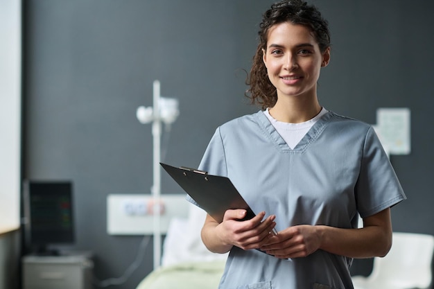 Retrato de jovem enfermeira com cartão médico sorrindo para a câmera em pé na enfermaria