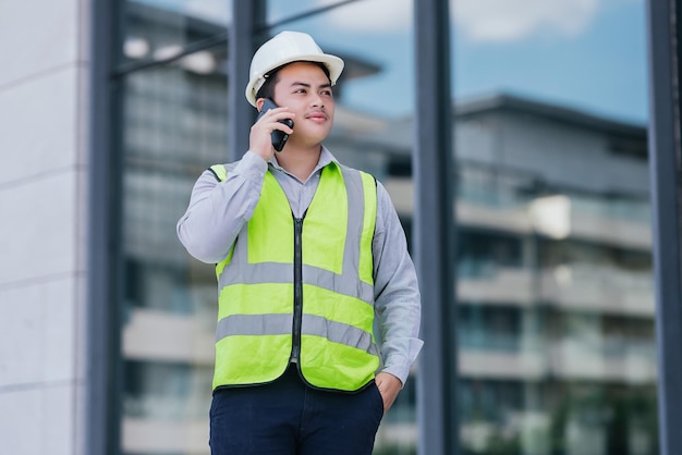 Retrato de jovem engenheiro asiático vestindo colete de segurança e capacete em pé usar smartphone para trabalho de contato no fundo do canteiro de obras Conceito de trabalhador da construção civil