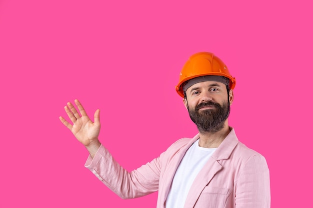 Retrato de jovem engenheiro de construção usa capacete laranja em uma jaqueta rosa em pé no fundo do estúdio vermelho O homem aponta com a mão