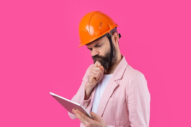 Retrato de jovem engenheiro de construção usa capacete laranja em uma jaqueta rosa em pé no fundo do estúdio vermelho Um homem com um tablet nas mãos