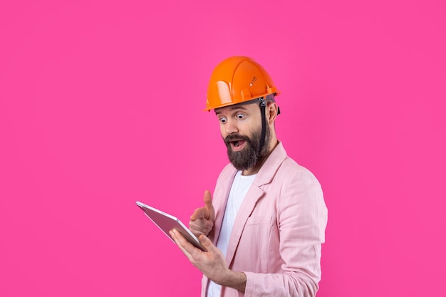 Retrato de jovem engenheiro de construção usa capacete laranja em uma jaqueta rosa em pé no fundo do estúdio vermelho Um homem com um tablet nas mãos