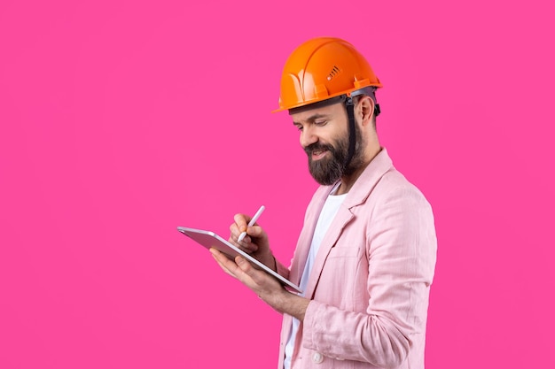 Retrato de jovem engenheiro de construção usa capacete laranja em uma jaqueta rosa em pé no fundo do estúdio vermelho Um homem com um tablet nas mãos