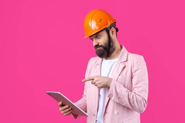 Retrato de jovem engenheiro de construção usa capacete laranja em uma jaqueta rosa em pé no fundo do estúdio vermelho Um homem com um tablet nas mãos