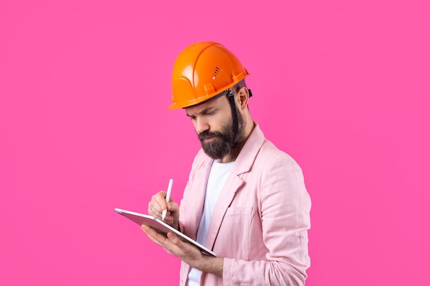 Retrato de jovem engenheiro de construção usa capacete laranja em uma jaqueta rosa em pé no fundo do estúdio vermelho Um homem com um tablet nas mãos