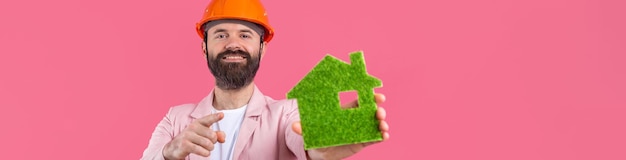Retrato de jovem engenheiro de construção usa capacete laranja em uma jaqueta rosa em pé no fundo do estúdio vermelho Um homem segura uma casa ecológica verde