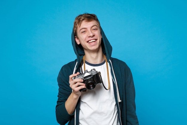 Retrato de jovem engraçado alegre em roupas casuais, segurando em mãos câmera fotográfica vintage retrô isolada na parede azul. Emoções sinceras de pessoas, conceito de estilo de vida.