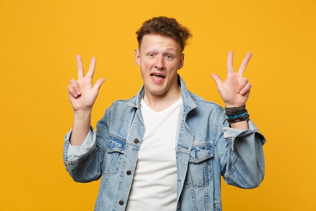 Retrato de jovem engraçado em roupas casuais jeans, olhando para a câmera, mostrando o sinal de vitória isolado em fundo amarelo laranja no estúdio. Emoções sinceras de pessoas, conceito de estilo de vida. Mock up espaço de cópia.