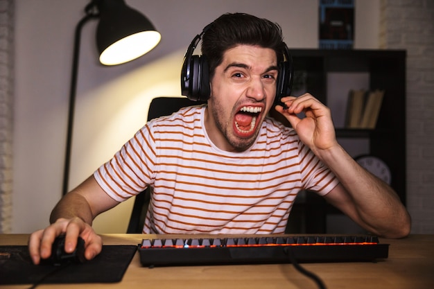 Retrato de jovem estressado de 20 anos usando fone de ouvido, gritando enquanto está sentado na mesa na sala e olhando para o monitor do computador