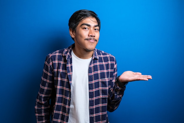 Retrato de jovem estudante asiático vestindo camisa, sorrindo e apontando para apresentar algo ao seu lado, contra um fundo azul com espaço de cópia