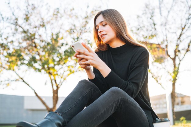 Retrato de jovem estudante com cabelos longos usando telefone celular no parque da cidade na hora de ouro