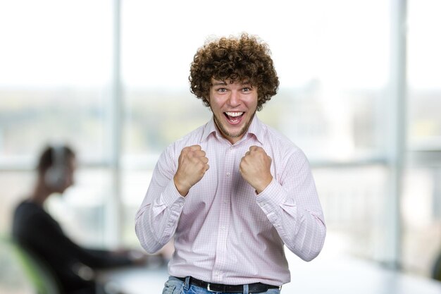 Retrato de jovem feliz e entusiasmado com cabelos encaracolados celebra o sucesso do interior do escritório interno no