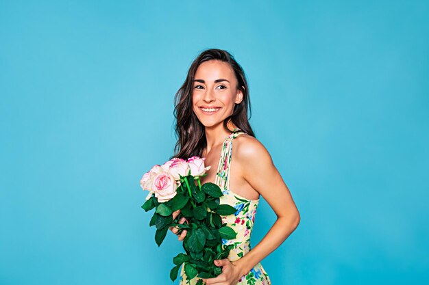 Retrato de jovem feliz e sorridente com cabelo longo encaracolado em vestido de verão com buquê de flores nas mãos posando sobre fundo azul Conceito de férias e presentes