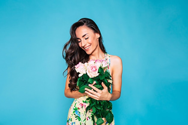 Retrato de jovem feliz e sorridente com cabelo longo encaracolado em vestido de verão com buquê de flores nas mãos posando sobre fundo azul conceito de férias e presentes