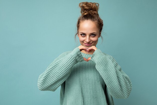 Retrato de jovem feliz positivo sorridente linda mulher loira com emoções sinceras, vestindo blusa azul casual isolada em um fundo azul com espaço de cópia e gesticulando com formato de coração de mãos