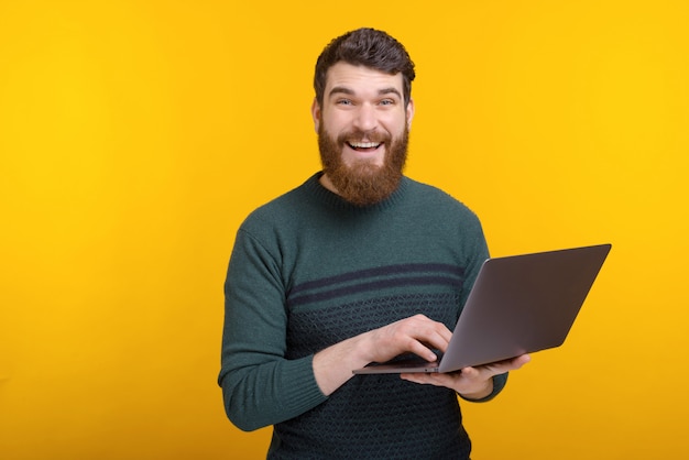 Retrato de jovem feliz usando laptop em pé sobre parede amarela