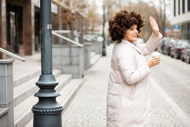 Retrato de jovem, fofa mulher caucasiana, 40 anos, esporte roupas casuais, andando acenando com a mão, xícara de café descartável. Penteado Afro. Vida na cidade, estilo de rua