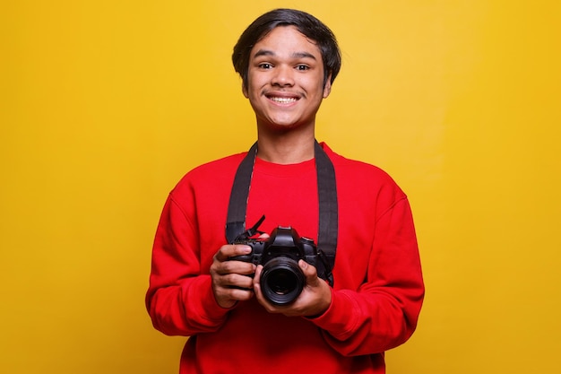 Retrato de jovem fotógrafo em estilo casual sorrindo enquanto segura a câmera contra um fundo amarelo