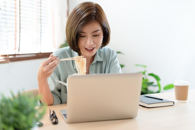 Retrato de jovem freelancer asiática trabalhando com laptop em sua casa e comendo comida asiática com comida para viagem Trabalho em casa durante a quarentena de auto-isolamento e Novo conceito normal